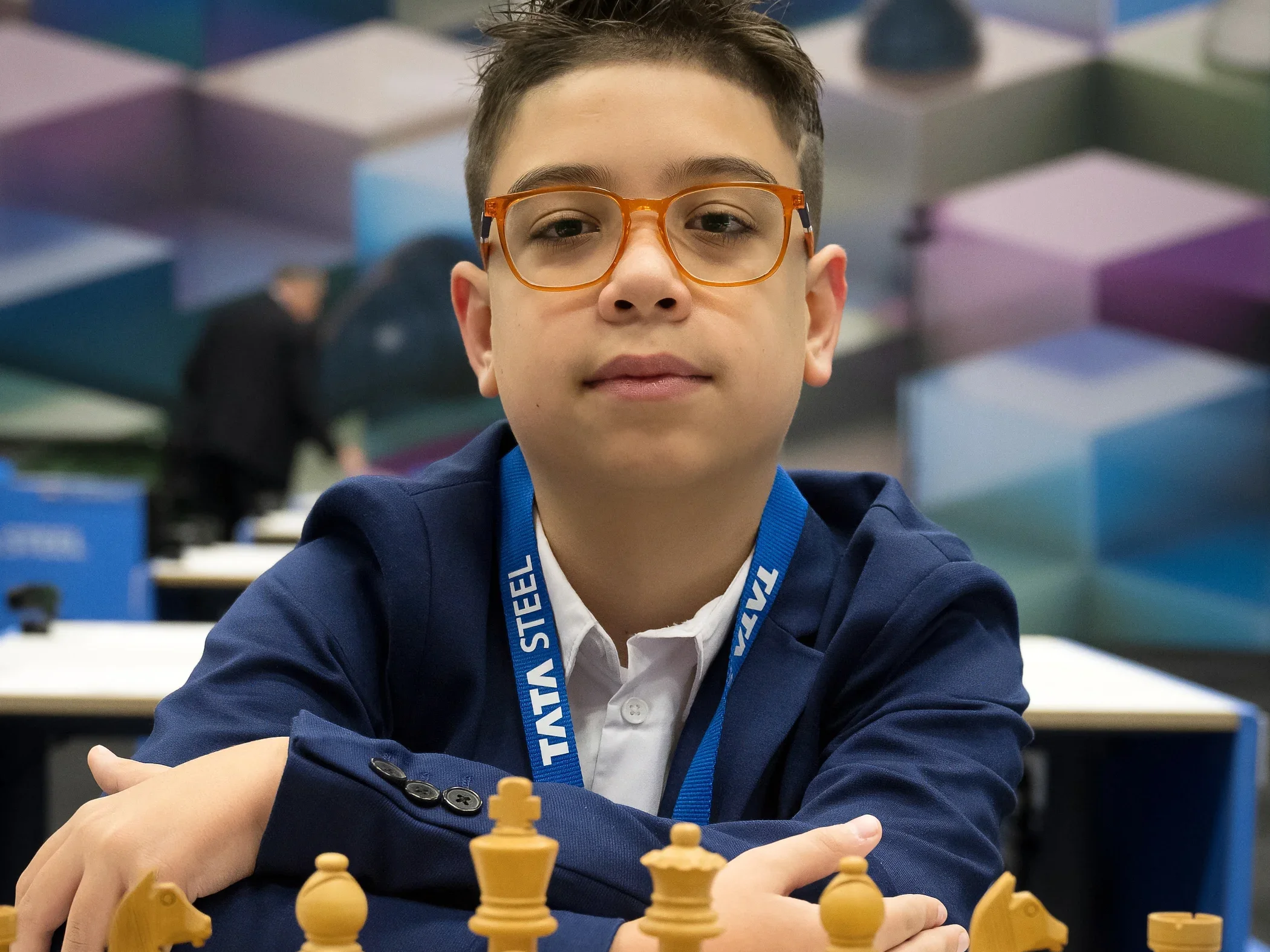 Faustino Oro seated at a chessboard at the 2025 Tata Steel tournament, wearing orange glasses and a tournament lanyard