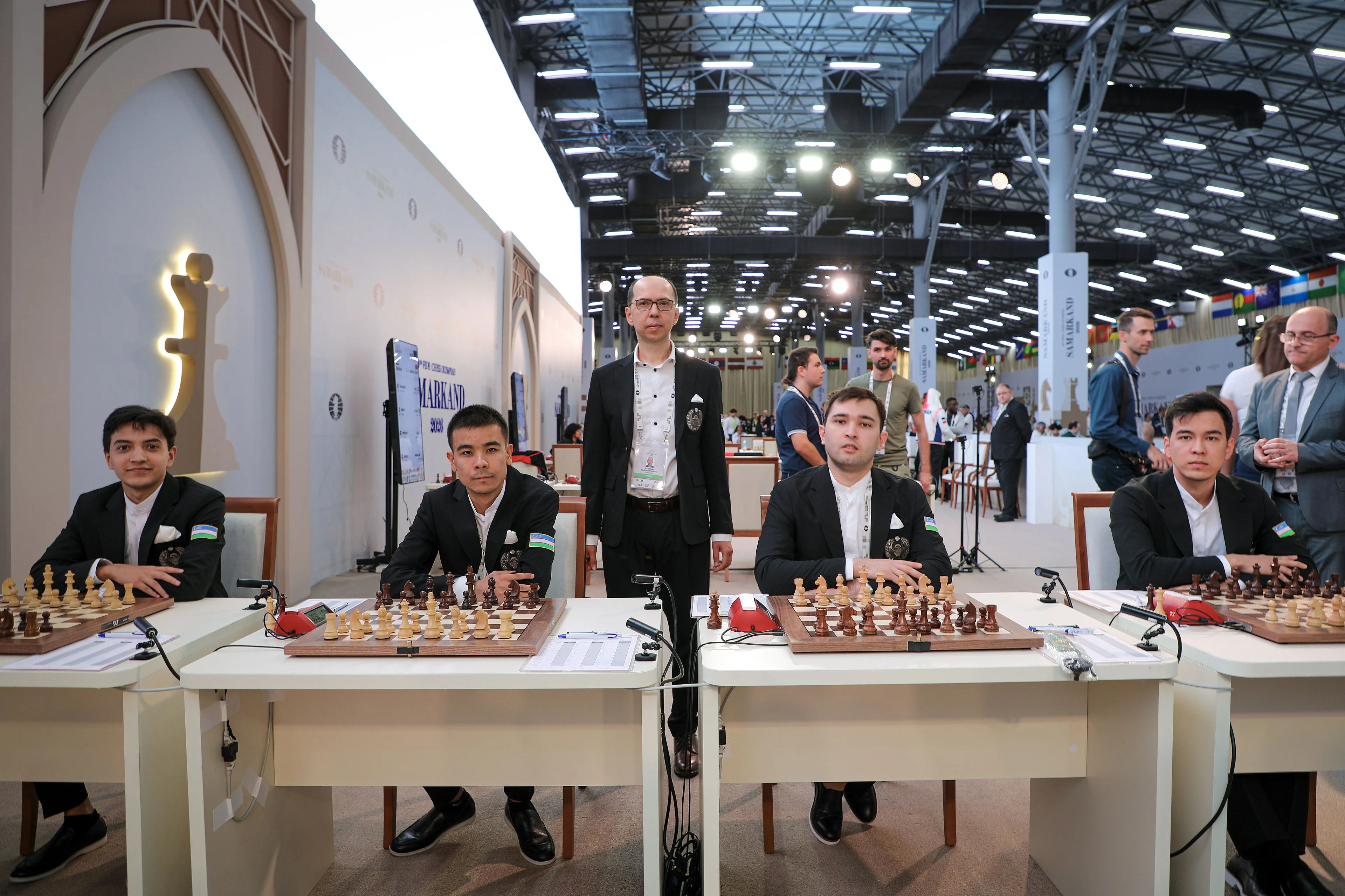 The Uzbekistan open team seated at their boards at the 46th Chess Olympiad in Samarkand, with their captain standing behind them
